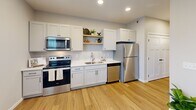 This efficiently designed one-bedroom, one-bathroom unit offers a thoughtfully organized layout across a single floor, maximizing every square foot of available space. The open-concept kitchen and living area serves as the heart of the unit, where light hardwood-style flooring, recessed lighting, and large windows create a bright and welcoming atmosphere. The kitchen is outfitted with white shaker cabinetry, white quartz countertops, stainless steel appliances including a range, microwave, dishwasher, and refrigerator, as well as open shelving for added storage and display. Adjacent to the kitchen, the living area provides a comfortable gathering space with natural light streaming through a large window fitted with blinds. A hallway equipped with a modern sliding barn door connects the main living space to the private areas of the unit. The bedroom, finished with plush carpet and neutral tones, offers a calm retreat. The full bathroom features wood-look tile flooring, a white vanity with ample storage, matte black hardware accents, and a tub-shower combination. Throughout the unit, a consistent neutral palette and clean, modern finishes contribute to a cohesive and functional living environment.