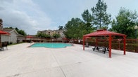 This inviting outdoor amenity space centers around a generously sized swimming pool with a freeform design, offering a refreshing retreat for residents. The pool is complemented by a broad concrete deck that provides ample room for lounging and socializing. A standout feature of the space is a covered gazebo with a bold red roof, furnished with a picnic table and seating, offering a shaded gathering spot ideal for outdoor dining or relaxation. Red accent umbrellas echo the gazebo's color scheme, adding a cohesive and vibrant aesthetic throughout the area. The space is enclosed by a perimeter fence, ensuring privacy and safety, while mature pine trees and leafy deciduous trees line the surroundings, creating a natural backdrop that softens the built environment. The pool area benefits from open sky exposure, allowing for abundant natural light throughout the day. In the background, the multi-story residential buildings are visible, providing context to the community setting. Overall, the space combines functional amenities with an attractive, well-maintained environment that encourages outdoor living.