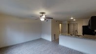 This single-story Dallas space offers a straightforward, functional layout designed for comfortable everyday living. The main living area is open and airy, finished with light-toned wood-look flooring that runs throughout, lending a clean and cohesive appearance to the space. A ceiling fan with an integrated light fixture is centrally positioned overhead, providing both air circulation and ambient lighting to the room. The walls are painted in a neutral warm beige, complementing the light flooring and creating a bright, welcoming atmosphere.

The kitchen is partially open to the living area, separated by a peninsula counter finished with a granite-style surface. This counter serves as both a workspace and a casual dining or gathering spot. Recessed lighting in the kitchen area ensures adequate task lighting. Cabinetry visible in the kitchen features a classic panel-door design in a warm wood tone. A doorway visible from the main living space leads further into the unit, suggesting additional rooms beyond. The overall design is clean and neutral, offering a versatile canvas well-suited to a variety of interior styles.