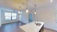 This single-story space showcases an open-concept layout that seamlessly connects the kitchen and living areas. The kitchen centers around a spacious island topped with a crisp white countertop and fitted with a stainless steel sink and contemporary gooseneck faucet. Dark cabinetry beneath the island provides a striking contrast to the light surfaces above. Three pendant lights hang above the island — a mix of cone and bell-shaped fixtures — adding both function and visual interest to the space.

The adjoining living area features hardwood-style flooring that extends throughout, contributing to a cohesive and polished aesthetic. A ceiling fan with a chandelier-style light fixture anchors the living space, while multiple windows allow natural light to filter in, creating a bright and welcoming atmosphere. A doorway visible from the kitchen leads to an additional room, suggesting a flexible floor plan suited for various uses. White walls and clean lines throughout maintain a modern, neutral palette, making the space feel open, airy, and move-in ready.