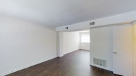 This two-story townhome offers a practical and well-organized layout across two floors, providing comfortable living with 2 bedrooms and 1.5 bathrooms. The main level features an open-concept living and dining area finished with rich, dark hardwood-style flooring that contrasts cleanly against crisp white walls and ceilings. A structural beam detail adds architectural character to the open living space, which flows naturally into a secondary area brightened by a large window that invites natural light. The entry foyer is welcoming, featuring a classic paneled front door with glass accent lights and direct sightlines into the main living area. A staircase with black metal railings leads to the upper floor, where the bedrooms and full bathroom are located. Conveniently situated near the entry, a louvered-door closet provides dedicated storage space. The kitchen area is positioned adjacent to the main living space, with a full-size refrigerator visible, offering practical functionality. A half-bathroom on the main level adds convenience for residents and guests alike. Throughout the home, HVAC vents and a wall-mounted heating unit ensure year-round comfort. The overall aesthetic is clean, neutral, and move-in ready.