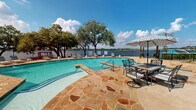 This two-story waterfront community is set along the shores of a expansive lake, offering residents direct access to the water and a range of thoughtfully designed outdoor amenities. The exterior features classic stone and siding architecture with covered balconies, chimneys, and large windows that frame the surrounding landscape. Winding concrete pathways connect the buildings to the community's recreational areas, passing through neatly manicured green lawns lined with mature trees.

At the heart of the outdoor space is a generously sized swimming pool surrounded by a distinctive flagstone patio deck. Wrought iron outdoor dining sets and shaded umbrella seating provide comfortable gathering areas poolside, all set against an unobstructed view of the lake just beyond the perimeter fence. Sun loungers line the pool area, making it an ideal spot for relaxation.

Descending toward the water, a private sandy beach area awaits, equipped with shade sail structures and teal lounge chairs. Stone-edged terracing connects the upper lawn to the shoreline, where the natural sandy beach meets the calm lake waters. The open waterfront offers panoramic views across the lake, completing the resort-like atmosphere of this lakeside community.