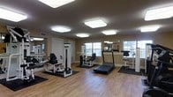 This fitness center offers a clean, functional environment designed to support a well-rounded workout routine. The space features warm hardwood flooring throughout, providing a polished and inviting atmosphere that complements the room's neutral beige walls and white trim. Overhead, a series of large, flush-mounted square ceiling lights ensure the entire area is evenly and brightly illuminated, enhancing visibility and creating an energizing workout environment. Multiple windows along the walls allow natural light to filter in, adding to the open and airy feel of the space. The room is outfitted with a thoughtful selection of exercise equipment, including strength-training machines such as a leg press and cable resistance units, as well as cardio equipment including a treadmill. Each piece of equipment is positioned on protective rubber floor mats, keeping the hardwood surface in excellent condition. Large wall mirrors are strategically placed to allow users to monitor their form during exercise and to visually expand the sense of space. A door provides access to an adjacent area, suggesting convenient connectivity to other amenities. Overall, the fitness center strikes a balance between practicality and comfort, making it a welcoming space for daily exercise.