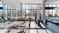 This expansive fitness center offers a generously sized, single-floor layout designed to accommodate a full range of workout activities. The space is defined by soaring ceilings, crisp white walls, and large floor-to-ceiling windows that flood the interior with natural light while providing open views of the surrounding urban streetscape. Structural columns accent the open floor plan, adding an architectural character to the contemporary design.

The facility is comprehensively equipped for both strength training and cardiovascular exercise. The strength zone features a wide array of Cybex Prestige selectorized machines, free weights with mirrored walls, and cable systems, all arranged to allow comfortable circulation between stations. The cardio area, positioned along the window wall to take full advantage of natural light and city views, houses multiple treadmills, elliptical trainers, stationary bikes, and rowing machines. An additional dedicated section offers stability balls, resistance accessories, and functional training equipment.

Durable dark rubberized flooring covers the workout areas, providing both safety and a sleek aesthetic contrast to the bright walls and ceilings. Mounted flat-screen televisions are positioned throughout the space for entertainment during workouts. The overall ambiance is clean, bright, and professional, reflecting a well-appointed fitness facility suited to a broad range of fitness needs.