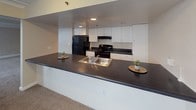 This single-floor residence offers a well-organized layout designed for comfortable everyday living. The open-concept kitchen serves as a functional hub, featuring a dark laminate peninsula countertop with a stainless steel sink, a full suite of black appliances including a refrigerator and electric range, and an abundance of white cabinetry providing generous storage. The kitchen flows naturally into the carpeted living areas, which are spacious and bright, benefiting from multiple large windows dressed with horizontal blinds that allow ample natural light to filter in.

Crown molding runs throughout the space, adding a refined architectural detail to the otherwise clean, neutral-toned walls. The flooring throughout the living and sleeping areas consists of wall-to-wall carpet in a warm beige tone, contributing to a cozy and quiet atmosphere. Several rooms feature built-in closets with sliding or bifold doors, offering practical storage solutions. The entry area is clearly defined with a paneled front door and open wall space ideal for customization.

The bathroom is equipped with a full-size soaking tub and shower combination, a vanity with a countertop sink, a large mirror, and wood-look flooring. The overall layout, as reflected in the 3D floorplan, is elongated and thoughtfully divided into distinct living, sleeping, and functional zones.