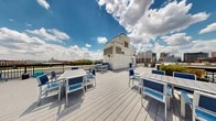 Located at 623 S Bethel St in Baltimore's vibrant Canton neighborhood, this rooftop terrace offers an impressive open-air retreat with sweeping panoramic views of the Baltimore cityscape. The deck is finished with light gray composite decking that spans a well-proportioned outdoor area, providing a clean and modern aesthetic underfoot.

The terrace is furnished with multiple seating arrangements, including white-framed dining tables paired with blue accent chairs — ideal for casual gatherings or al fresco dining. A separate lounge area features plush wicker sectional seating, offering a more relaxed setting to take in the skyline. Raised planter boxes with manicured greenery add a touch of natural contrast against the urban backdrop, softening the rooftop environment.

A secure metal railing encircles the perimeter, ensuring safety while preserving unobstructed sightlines across the Baltimore skyline in all directions. The rooftop is accessed via a dedicated elevator lobby finished with slate tile flooring, stainless steel elevator doors, and neutral white walls — presenting a polished and functional transition point between the building interior and the outdoor terrace. Natural light fills the elevator lobby through a large window. Together, these elements make this rooftop terrace a standout communal amenity with impressive urban views.