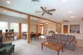 This single-floor Cedar facility is thoughtfully designed to accommodate a variety of uses, combining comfort and functionality throughout its well-organized layout. The centerpiece of the space is a generously sized lounge area featuring warm hardwood-style flooring, a drop panel ceiling with recessed lighting, and multiple large windows and glass doors that invite ample natural light and provide views to the outdoors. Comfortable upholstered sofas and loveseats are arranged around a central coffee table atop a patterned area rug, creating an inviting gathering atmosphere. A wall-mounted flat-screen television and a stone-faced fireplace surround add character and entertainment value to the lounge. Structural wood beam accents lend a warm, lodge-inspired aesthetic throughout. A wet bar or lounge counter area is visible in the background, adding convenience for events or social gatherings. The fully equipped kitchen features warm oak cabinetry, granite-style countertops, and black appliances including a range, over-the-range microwave, and dishwasher. A clean entry corridor with branded Cedar floor mats leads to restroom facilities and additional rooms accessible via interior doors, with a staircase providing access to other levels of the building.