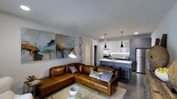 This single-floor space showcases a thoughtfully designed open-concept layout that seamlessly connects the living area and kitchen into one cohesive environment. The living area is anchored by a rich cognac leather sectional sofa accented with decorative pillows, positioned atop a textured area rug over polished concrete floors. A glass coffee table completes the seating arrangement, while large abstract canvases in blue, gold, and white add a vibrant artistic focal point to the white walls. Eclectic industrial-style pendant and floor lamps contribute a layered, curated lighting scheme throughout the space.

The kitchen flows naturally from the living area and features two-tone cabinetry with light upper cabinets and darker lower units, topped with a light stone countertop that extends into a breakfast bar with bar-stool seating. Stainless steel appliances, including a refrigerator and range, are complemented by a subway tile backsplash. Sleek black pendant lights hang above the peninsula, reinforcing the contemporary industrial aesthetic that defines the overall ambiance of this well-appointed space.
