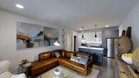 This single-floor space showcases a thoughtfully designed open-concept layout that seamlessly connects the living area and kitchen into one cohesive environment. The living area is anchored by a rich cognac leather sectional sofa accented with decorative pillows, positioned atop a textured area rug over polished concrete floors. A glass coffee table completes the seating arrangement, while large abstract canvases in blue, gold, and white add a vibrant artistic focal point to the white walls. Eclectic industrial-style pendant and floor lamps contribute a layered, curated lighting scheme throughout the space.

The kitchen flows naturally from the living area and features two-tone cabinetry with light upper cabinets and darker lower units, topped with a light stone countertop that extends into a breakfast bar with bar-stool seating. Stainless steel appliances, including a refrigerator and range, are complemented by a subway tile backsplash. Sleek black pendant lights hang above the peninsula, reinforcing the contemporary industrial aesthetic that defines the overall ambiance of this well-appointed space.