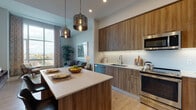 This single-floor residence combines contemporary design with functional living across an open, well-organized layout. The kitchen is a standout feature, outfitted with warm wood-grain cabinetry extending from floor to ceiling, sleek white quartz countertops, a hexagonal tile backsplash, and a full suite of stainless steel appliances including a range, built-in microwave, dishwasher, and refrigerator. A central island provides additional prep space and casual dining, while a pair of brushed metallic pendant lights adds a refined industrial touch. Light wood flooring runs throughout, lending warmth and continuity to the space.

The open-concept living area benefits from dramatic floor-to-ceiling windows that frame expansive city views, flooding the room with natural light. Neutral walls and generous ceiling height contribute to an airy, elevated atmosphere.

The bedroom continues the light-toned palette, featuring large windows with city views, plush carpeting, and ample wall space. The bathroom is finished with a wood-front vanity, white quartz countertop, light tile flooring, and a tub-shower combination accented by deep charcoal subway tile, creating a clean, contemporary aesthetic throughout.