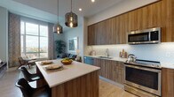 This single-floor residence combines contemporary design with functional living across an open, well-organized layout. The kitchen is a standout feature, outfitted with warm wood-grain cabinetry extending from floor to ceiling, sleek white quartz countertops, a hexagonal tile backsplash, and a full suite of stainless steel appliances including a range, built-in microwave, dishwasher, and refrigerator. A central island provides additional prep space and casual dining, while a pair of brushed metallic pendant lights adds a refined industrial touch. Light wood flooring runs throughout, lending warmth and continuity to the space.

The open-concept living area benefits from dramatic floor-to-ceiling windows that frame expansive city views, flooding the room with natural light. Neutral walls and generous ceiling height contribute to an airy, elevated atmosphere.

The bedroom continues the light-toned palette, featuring large windows with city views, plush carpeting, and ample wall space. The bathroom is finished with a wood-front vanity, white quartz countertop, light tile flooring, and a tub-shower combination accented by deep charcoal subway tile, creating a clean, contemporary aesthetic throughout.