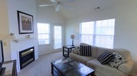 This single-floor residence centers around a well-appointed living room that immediately draws attention with its vaulted ceiling, which adds a sense of openness and grandeur to the space. A ceiling fan is mounted at the peak, providing both functional air circulation and a finishing architectural touch.

Natural light fills the room through multiple large windows fitted with white horizontal blinds, allowing for adjustable brightness throughout the day. A glass-paneled door provides direct access to an outdoor area, further enhancing the airy, connected feel of the interior.

The living room is finished with neutral wall tones and plush wall-to-wall carpeting, creating a warm and comfortable atmosphere. Furnishings include a beige sectional sofa accented with patterned throw pillows, dark wood side and coffee tables with glass insets, and a table lamp that adds a soft, ambient glow. A fireplace is visible along one wall, offering an additional focal point and a cozy element to the overall space.

The layout is thoughtfully arranged to maximize both comfort and functionality, making this a practical and inviting living environment.