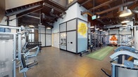 The weight room at 200 Redpath Avenue is a two-storey fitness facility with a striking industrial aesthetic. Soaring double-height ceilings on the main floor are framed by floor-to-ceiling windows that flood the space with natural light and offer panoramic views of the Toronto skyline. Exposed ductwork, dark steel accents, and grid-patterned partition walls contribute to the facility's urban, warehouse-inspired character.

The main floor houses an extensive selection of strength training equipment, including a full dumbbell rack, adjustable benches, cable machines, and functional training stations. A branded functional training zone — "The Temple" — features a multi-rig system, battle ropes, a heavy bag, resistance equipment, and an artificial turf runway. A water station and change room access are conveniently located on this level.

A steel staircase connects to the upper floor, where a dedicated spin studio features hardwood flooring, full-wall mirrors, and large windows with city views. Treadmills positioned directly against the expansive glazing allow users to work out while overlooking the Toronto cityscape. Industrial pendant lighting completes the polished yet functional atmosphere throughout both levels.