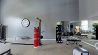 Located at 3920 Knickerbocker Pkwy in Raleigh, NC, this fitness center offers a well-appointed, single-floor workout facility designed to support a wide range of exercise routines. The space is finished with neutral gray walls and low-pile carpet flooring, creating a clean and focused environment conducive to training.

The gym is equipped with a notable variety of fitness equipment. A prominent heavy bag and multi-functional rack system anchors one section of the space, offering options for boxing and strength training. A dedicated storage tower holds an assortment of kettlebells and medicine balls in multiple weights, making circuit and functional training readily accessible. A weight bench and additional free weights complement the strength training setup.

Full-length mirrors line one wall, visually expanding the space and allowing users to monitor their form during workouts. The mirrored section also reflects cardio equipment positioned toward the back of the room, including what appears to be a treadmill. A large decorative wall clock adds a practical and stylish touch to the overall aesthetic.

The layout is thoughtfully organized to provide ample floor space for movement-based exercises, stretching, or battle rope training. Overall, the fitness center delivers a comprehensive and functional workout environment suitable for varied fitness goals.