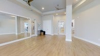 This yoga studio offers a welcoming and functional space thoughtfully designed to support movement and mindfulness. The main practice area is generously proportioned, featuring smooth, light-toned hardwood flooring that runs throughout, providing a clean and stable surface ideal for yoga and other wellness activities. Elevated ceilings with recessed lighting and dual ceiling fans create an airy, well-ventilated atmosphere that contributes to a comfortable practice environment regardless of the season.

A large mirrored wall panel along one side enhances the sense of openness while allowing practitioners to observe and refine their form during sessions. The neutral, soft gray and white color palette throughout maintains a serene and distraction-free ambiance conducive to focused practice.

The studio includes multiple doorways leading to additional rooms, which appear to offer supplementary spaces such as changing areas or ancillary rooms. White-framed glass panel doors allow natural light to filter between spaces while maintaining a cohesive, clean aesthetic. A structural column is integrated discreetly into the layout without significantly interrupting the open floor plan. Overall, the studio balances practicality with a calm, minimalist design sensibility.