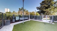 This thoughtfully designed outdoor pet relief area provides a clean, functional, and welcoming environment for pets and their owners. The space is surfaced with lush artificial turf, offering a hygienic and low-maintenance ground cover that stays green year-round. A secure perimeter of white metal mesh fencing encloses the area, ensuring the safety of pets while maintaining an open, airy feel. Overhead, a metal pergola-style structure adds architectural definition and frames the sky above. A pair of metal benches lines one side of the enclosure, giving pet owners a comfortable place to sit while their companions enjoy the space. Landscape lighting poles are positioned throughout, suggesting the area is fully usable during evening hours as well. An informational station with signage and a waste disposal unit is conveniently located near the entrance, promoting cleanliness and responsible pet ownership. The surrounding landscape features mature palm trees and dense green foliage, creating a natural tropical backdrop that enhances the overall ambiance. The combination of practical amenities, secure boundaries, and verdant surroundings makes this a well-appointed and inviting outdoor destination for pets.