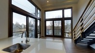 This contemporary two-story space is defined by its striking architectural design and abundant natural light. The main living area showcases dramatic floor-to-ceiling windows with bold dark frames, drawing in sweeping views of the surrounding neighborhood and mature trees. The generous ceiling height amplifies the sense of openness, creating an airy, expansive atmosphere throughout.

The kitchen features a sleek, light-colored countertop with a modern undermount sink and a polished faucet, reflecting a clean and functional aesthetic. The open-concept layout allows the kitchen to flow seamlessly into the main living area, making the space ideal for both daily living and entertaining.

Warm hardwood flooring runs throughout the main level, adding a natural contrast to the contemporary dark window frames and modern finishes. Recessed lighting complements the natural light that pours in from the large windows. A staircase connects the two floors, adding to the vertical dimension and architectural interest of the space.

Overall, the space strikes a balance between modern design sensibility and warm, livable comfort, with its expansive windows, high ceilings, and thoughtfully chosen materials creating a bright, welcoming environment with strong visual character.
