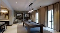 This single-floor space is designed with entertainment and relaxation in mind, blending modern aesthetics with warm, inviting elements. At the heart of the room sits a full-size billiards table finished in dark wood and charcoal felt, positioned beneath a striking industrial-style chandelier with exposed Edison bulbs that serves as a bold focal point. The vaulted ceiling adds an impressive sense of height and openness to the space.

A dramatic stacked stone accent wall anchors the lounge area, featuring built-in niches with framed artwork and integrated lighting that highlights the rich, textured surface. Comfortable seating — including upholstered chairs and a cushioned bench — is thoughtfully arranged to create an intimate gathering area. Neutral tones throughout, from the soft beige walls to the plush area rug, complement the darker furnishings and stone features.

Natural light filters in through floor-to-ceiling windows dressed with flowing linen drapes, adding an airy, elegant contrast to the room's moody, sophisticated palette. Recessed ceiling lighting provides additional ambient illumination. The overall ambiance is refined yet inviting — a well-appointed space ideal for socializing and leisure.