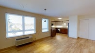 This single-floor unit features an open-concept layout that seamlessly connects the living area to the kitchen, creating a cohesive and functional living space. Light hardwood flooring extends throughout, lending warmth and visual continuity across the entire floor plan.

The living area is bright and airy, benefiting from two large windows fitted with horizontal blinds that allow for adjustable natural light. A wall-mounted HVAC unit sits beneath the windows, providing climate control. The warm beige walls complement the flooring and contribute to the overall neutral palette of the space.

The kitchen is well-appointed with a two-tone cabinet design, combining rich dark wood lower cabinets with lighter upper cabinetry topped by dark accents. A central island with a pendant light overhead provides additional workspace and a casual gathering point. Stainless steel appliances, including a refrigerator, dishwasher, and microwave, are integrated throughout. A sink is positioned along the back wall beneath under-cabinet lighting, adding both functionality and ambiance. The kitchen's efficient layout makes good use of the available space while maintaining an open, accessible feel.