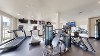 This fitness center is a thoughtfully designed, single-level facility offering a comprehensive range of workout equipment to support diverse training needs. The space is divided into distinct functional zones, each tailored to a specific type of exercise.

The cardio area features multiple treadmills and other aerobic machines positioned along windowed walls, allowing users to enjoy natural light and views of the surrounding outdoor landscaping while exercising. Wall-mounted flat-screen televisions are positioned throughout, providing entertainment during workouts.

The strength training section includes a multi-functional cable machine, a variety of selectorized weight machines, and a dedicated free weights area equipped with a full rack of dumbbells and adjustable benches. Full-length mirrors line portions of the walls, enhancing the sense of space and allowing users to monitor their form.

Flooring transitions between rubberized material in high-use equipment zones and smoother surfaces in open areas, offering both durability and functionality. Recessed lighting supplements the abundant natural light entering through large windows and glass doors along the perimeter. A vending machine is also present, providing convenient access to refreshments. The overall ambiance is clean, modern, and well-maintained, creating a practical and comfortable environment for fitness activities.