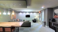 This contemporary single-floor space is thoughtfully designed to serve as a comfortable and functional communal lounge. The open layout features a well-appointed seating area anchored by a plush gray sectional sofa accented with warm-toned throw pillows, complemented by a sleek black accent chair with a tripod floor lamp nearby. A wall-mounted flat-screen television serves as a focal point for the lounge area, making it ideal for both relaxed gatherings and entertainment.

The interior is finished with neutral gray walls and patterned low-pile carpeting that adds texture underfoot while maintaining a clean, modern aesthetic. Recessed ceiling lighting provides bright, even illumination throughout the space, enhanced by stylish pendant lights that hang near a live-edge wood communal table, suggesting a seamlessly integrated dining or collaborative work area.

The ceiling incorporates subtle architectural detailing with contrasting dark panels that add visual depth and dimension. A door with a sidelight panel allows natural light to filter into the space, connecting the interior to an adjacent exterior area. Abstract wall art adds a pop of color, reinforcing the modern, welcoming atmosphere of this well-designed lounge space.