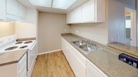 This single-floor unit offers a practical and functional layout with a blend of neutral tones and varied flooring throughout. The living area is generously proportioned, featuring plush wall-to-wall carpeting and large vertical-blind-covered sliding glass doors that open to a balcony, allowing natural light to fill the space. A wall-mounted HVAC unit ensures year-round climate comfort in the living area.

The kitchen is designed in a galley style, featuring white cabinetry with ample upper and lower storage, speckled stone-look countertops, a double stainless steel sink, and a skylight overhead that brightens the workspace. Wood-look flooring runs through the kitchen and adjoining dining or entry area, creating a clean transition between spaces. A breakfast bar connects the kitchen to the main living area, enhancing the open flow.

The bedroom is well-sized with soft carpeting, neutral walls, and a wide horizontal window fitted with vertical blinds for light control and privacy.

The bathroom features an extended vanity with dual sinks, white cabinetry, stone-look countertops, a recessed medicine cabinet, and a separate room housing a full tub-shower combination. Textured ceilings are present throughout the unit, adding a classic finishing detail.