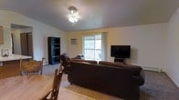 Located at 515-535 W Schindler Pl in Menasha, Wisconsin, this single-floor residence offers a practical and comfortable layout designed for everyday living. The main living area serves as the heart of the space, featuring neutral-toned walls and carpeted flooring that contribute to a warm, understated ambiance. A ceiling fan with integrated lighting provides both air circulation and overhead illumination. The living room is generously sized, accommodating a large seating arrangement alongside additional storage and entertainment furnishings with ease. A sliding glass door floods the room with natural light and opens directly onto a private balcony with a railing, offering outdoor views of the surrounding greenery — a notable feature that enhances the overall sense of openness. A wall-mounted air conditioning unit provides supplemental climate control. The dining area is situated adjacent to the living space, with a wooden table and chairs providing a dedicated spot for meals. The single-floor layout ensures convenient, step-free access throughout, making the space both accessible and easy to navigate. Overall, the unit presents a functional and livable environment in a residential Menasha setting.