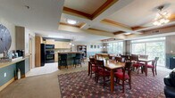 Located at 18351 Kenyon Ave in Lakeville, MN, this community club room offers a thoughtfully designed, multi-functional gathering space spread across a single floor. The layout encompasses a fully equipped kitchen, a dining area, a comfortable living room, and a dedicated billiards area, making it an ideal venue for social events and community activities.

The kitchen features light maple cabinetry, a tile backsplash, black appliances including a refrigerator, range, and microwave, and a granite-topped island with bar seating for casual dining. Pendant lighting and tile flooring complete the functional yet inviting look.

The dining area accommodates multiple tables with rich wood finishes and red-upholstered chairs, set atop a patterned area rug. Coffered ceilings with warm wood trim lend architectural distinction to this zone.

The living room offers a stone-surround fireplace with a wood mantel, leather seating, and a wall-mounted television, creating a cozy retreat. Tray ceilings with wood accents and striped wallpaper add a refined touch.

The billiards area centers on a full-size pool table beneath a statement hanging light fixture. Large windows flood the space with natural light and offer lush, tree-lined views, seamlessly connecting the interior to the surrounding landscape.