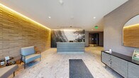 The 2nd Floor Lobby at The Mill presents a refined and contemporary welcome experience, thoughtfully designed to balance functionality with aesthetic appeal. Upon entry through expansive floor-to-ceiling glass doors, visitors are greeted by polished terrazzo-style stone flooring that extends throughout the space, lending a sense of elegance and solidity underfoot.

At the heart of the lobby stands a sleek, modern reception desk framed by a dramatic large-scale photographic mural depicting a feather and crashing wave motif, serving as a bold artistic focal point. Pendant lighting suspended above the desk adds a refined finishing touch. To one side, a textured stacked-stone accent wall bathed in warm amber cove lighting introduces a natural, organic contrast to the otherwise cool, neutral palette of the space.

Seating areas with contemporary upholstered chairs offer comfortable waiting areas for visitors, while a stylish chevron-patterned console adds a decorative accent near the entrance. A mailroom alcove is tucked conveniently behind the mural wall. The elevator lobby, finished in layered gray textured wallcovering with matching tile flooring and a minimalist bench, provides a calm and composed transition to upper floors. The overall ambiance is polished, professional, and welcoming.