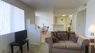This single-floor residence is designed with an open-concept floor plan that seamlessly connects the main living areas, creating a sense of spaciousness and ease of movement throughout. The living room serves as a central gathering space, carpeted for comfort and furnished with a plush sofa accented by multicolored throw pillows. A flat-screen television is positioned across from the seating area, with a large window on the adjacent wall allowing natural light to fill the room.

A low half-wall divider separates the living room from the dining area, which features hardwood-style flooring, a round dining table, and wicker-style chairs beneath a pendant light fixture. This transition from carpet to hard flooring helps define each zone while maintaining the open feel of the layout.

Beyond the dining area, the kitchen is equipped with light-toned cabinetry, a breakfast bar counter, overhead lighting, and standard appliances including a stove and refrigerator. The layout allows visibility across all three areas from nearly any vantage point, making the space feel cohesive and well-connected. The overall ambiance is casual, bright, and practical.