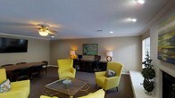 This single-floor space offers a thoughtfully designed interior that balances comfort and functionality. The main living and lounge area serves as the centerpiece, featuring a set of bold yellow accent chairs arranged around a glass-top coffee table with ornate gold detailing, creating a vibrant focal point against the neutral, taupe-toned walls. A textured popcorn ceiling adds a classic architectural touch, while a ceiling fan with an integrated light fixture ensures both air circulation and ambient lighting.

Crown molding runs along the upper perimeter of the room, lending a polished, finished look to the space. Recessed lighting complements the warm glow of matching table lamps flanking a dedicated workspace, which includes a dark wood desk and a framed map artwork as a decorative backdrop. The workspace area is well-lit and organized, making it suitable for both professional and personal use.

Dark carpeting anchors the room and provides a grounding contrast to the lighter wall tones and bright furnishings. A small dining or accent table with chairs is visible in the corner, adding to the room's versatility. Overall, the space presents a cohesive, comfortable environment with a mix of functional zones and curated décor.