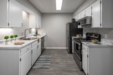 This single-floor residence offers a thoughtfully designed layout that balances functionality and comfort throughout its key living spaces. The galley-style kitchen is a standout feature, showcasing crisp white cabinetry, subway tile backsplash, and light marble-look countertops that run the length of both walls. A full suite of stainless steel appliances — including a refrigerator, electric range, over-the-range microwave, and dishwasher — equips the kitchen for everyday use. A large skylight-style overhead fixture provides bright, even illumination, while wood-look flooring adds warmth and continuity underfoot. Adjacent to the kitchen is an open dining or flex area, finished with the same wood-look flooring and neutral wall tones, offering versatile use of the space. The bedroom is finished with plush wall-to-wall carpeting, neutral walls, and a window fitted with blinds that allow natural light to filter in. A ceiling fan overhead provides year-round comfort. The bathroom features a white vanity with a marble-look countertop, undermount sink, and a large mirror, complemented by tile flooring and clean, modern finishes. Throughout the residence, a cohesive neutral color palette and consistent modern finishes create a clean and inviting atmosphere.