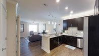 This single-floor residence showcases a thoughtfully designed open-concept layout that seamlessly connects the kitchen and living areas into one cohesive, functional space. The kitchen is a standout feature, outfitted with rich dark cabinetry contrasted by light quartz countertops and a subway tile backsplash. A peninsula with pendant lighting overhead provides additional prep space and casual seating, while a built-in wine cooler adds a refined touch. Recessed lighting throughout ensures the space remains bright and well-lit. The kitchen flows naturally into the spacious living area, which benefits from natural light streaming in through multiple windows. A ceiling fan adds comfort to the living space, which is finished with the same warm wood-style flooring found throughout the unit. Neutral white walls provide a clean, versatile backdrop, creating a bright and welcoming ambiance. The cohesive design elements — from the consistent flooring to the modern fixtures — give the entire space a polished, contemporary aesthetic well-suited for both everyday living and entertaining.