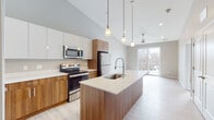 This single-floor residence showcases a thoughtfully designed open-concept layout that seamlessly connects the kitchen and living areas. The kitchen is a standout feature, combining two-tone cabinetry — warm walnut-finish lower cabinets paired with high-gloss white upper cabinets — atop light quartz countertops. A generously sized center island with an integrated sink and a professional-style pull-down faucet serves as both a functional workspace and a natural gathering point. Stainless steel appliances, including an electric range, over-the-range microwave, and French door refrigerator, complete the well-appointed cooking space. Pendant lighting suspended from elevated ceilings adds visual interest and draws the eye upward, enhancing the sense of openness throughout.

The living area extends naturally from the kitchen and is bathed in natural light from a large sliding glass door that opens to a private balcony, offering outdoor views of surrounding trees. Light wood-tone flooring runs throughout the main living spaces, while the bedroom features plush carpet and a large window with scenic views. The bathroom includes a full tub-shower combination, wood-toned vanity cabinetry, and clean neutral finishes, maintaining the modern aesthetic found throughout the space.