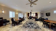 This single-floor space features an open-concept layout that seamlessly integrates the dining area, living lounge, and kitchen into one cohesive environment. The dining area is anchored by a warm wooden round table surrounded by dark ladder-back chairs, positioned near windows that allow natural light to filter in through shuttered panels. The living area centers around a striking glass accent table topped with a tall, ornate black vase, set atop a bold black-and-white damask area rug. Sleek black leather seating completes the lounge arrangement. Dual ceiling fans with brushed nickel detailing are mounted throughout, contributing to the airy feel of the space. The flooring throughout is a large-format natural stone tile in warm beige and tan tones, adding texture and sophistication. The kitchen, visible in the background, features rich dark wood cabinetry, stainless steel appliances, and a mosaic tile backsplash that serves as a standout design detail. Recessed lighting and wall sconces provide layered illumination, while crown molding along the ceiling adds a polished, refined finish to the overall space.