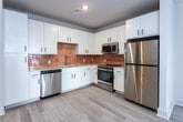 Located at 215 Willow Ave in Knoxville, Tennessee, this single-floor unit offers a clean, modern design throughout. Light-toned hardwood-style flooring runs continuously across the entire space, creating a cohesive and airy feel. The open-concept living and dining area flows seamlessly into the kitchen, which is a standout feature of the unit. The kitchen is outfitted with crisp white shaker-style cabinetry, a warm terracotta-toned tile backsplash, stainless steel appliances including a gas range, microwave, dishwasher, and refrigerator, making it both functional and visually striking. Natural light filters in through multiple windows, brightening each room. The bedroom includes a ceiling fan and its own window, while a generously sized walk-in closet with wire shelving provides ample storage. The bathroom features light wood-style flooring, a white vanity with a large mirror, and a soaking tub with full tile surround in an earthy stone finish. An in-unit washer and dryer are conveniently situated within the living area. The overall design palette of neutral whites and warm accents gives the space a fresh, move-in-ready character well-suited for comfortable everyday living.