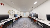 Located at 308 Scott St SW in Atlanta, Georgia, this single-room computer lab office is thoughtfully designed to support a productive and tech-forward work environment. The space is organized around two parallel rows of white marble-top workstations running the length of the room, each equipped with desktop computers and paired with sleek black and orange chairs that add a pop of color to the neutral palette.

The room's white walls are clean and bright, illuminated by recessed ceiling lighting that ensures consistent, glare-free coverage throughout. A standout design feature is the bold navy blue circuit board mural adorning one wall, reinforcing the technology-focused purpose of the space. Complementing this, two large decorative clocks — one with a Roman numeral face and one with a minimalist gold frame — are mounted on the opposite wall, adding character and visual interest.

A centrally positioned window with white blinds allows natural light to filter in, enhancing the open and airy feel of the room. Patterned gray carpet tiles cover the floor, adding texture while maintaining a professional aesthetic. Near the entry, a door with flanking windows provides transparency into adjacent spaces, and a printer station is conveniently accessible. The overall ambiance is clean, modern, and purpose-built for collaborative or individual computer-based work.