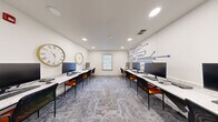 Located at 308 Scott St SW in Atlanta, Georgia, this single-room computer lab office is thoughtfully designed to support a productive and tech-forward work environment. The space is organized around two parallel rows of white marble-top workstations running the length of the room, each equipped with desktop computers and paired with sleek black and orange chairs that add a pop of color to the neutral palette.

The room's white walls are clean and bright, illuminated by recessed ceiling lighting that ensures consistent, glare-free coverage throughout. A standout design feature is the bold navy blue circuit board mural adorning one wall, reinforcing the technology-focused purpose of the space. Complementing this, two large decorative clocks — one with a Roman numeral face and one with a minimalist gold frame — are mounted on the opposite wall, adding character and visual interest.

A centrally positioned window with white blinds allows natural light to filter in, enhancing the open and airy feel of the room. Patterned gray carpet tiles cover the floor, adding texture while maintaining a professional aesthetic. Near the entry, a door with flanking windows provides transparency into adjacent spaces, and a printer station is conveniently accessible. The overall ambiance is clean, modern, and purpose-built for collaborative or individual computer-based work.