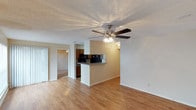 This single-floor residence in Denton offers a practical and comfortable layout designed for easy everyday living. The main living area is open and airy, finished with warm hardwood-style flooring that extends throughout the space, adding a cohesive and inviting feel. A ceiling fan with an integrated light fixture anchors the living room, providing both comfort and ambient lighting. The kitchen is positioned adjacent to the living area and features a raised breakfast bar that serves as a natural dividing point, along with dark-toned cabinetry, a black refrigerator, and a built-in countertop range, making it a functional space for meal preparation and casual dining. Neutral, light-toned walls throughout the home create a clean and versatile backdrop. A doorway from the living area leads to an additional room, visible in the background, suggesting a flexible layout that can accommodate a variety of needs. The textured ceiling adds a classic touch consistent with the overall character of the home. The space is well-suited for those seeking a straightforward, move-in-ready residence with a warm and functional interior in the Denton area.