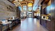 The Trails at Timberline clubhouse is a thoughtfully designed community amenity space that blends rustic mountain aesthetics with modern finishes throughout. The grand entry lobby immediately impresses with its soaring vaulted ceiling, exposed timber beam framework, reclaimed wood accent walls, and large pendant light fixtures that bathe the expansive tile flooring in warm light. Tall clerestory windows flood the space with natural light, while glass entry doors open to outdoor views.

The lounge area features a striking floor-to-ceiling stone fireplace flanked by oversized windows with dark wood framing, plush seating, and richly patterned carpeting that adds warmth and texture. Adjacent to the lounge, a fully equipped kitchen and bar area offers stainless steel appliances, granite countertops, dark cabinetry, pendant lighting, and bar stool seating — ideal for community gatherings.

A dedicated media room provides a true cinematic experience, complete with a large projection screen, padded wall panels, tiered soft seating, and bean bag chairs for a relaxed atmosphere. The games area includes a billiards table set on rich hardwood flooring, alongside a bowling alley with its own viewing seating and mounted display screens. Private office or meeting space rounds out the amenities, featuring carpeted flooring, natural light, and refined décor.