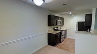 This single-floor space offers a practical and modern layout designed for efficient living. The kitchen serves as a functional centerpiece, featuring sleek dark espresso cabinetry that provides a striking contrast against the light-toned walls. Stainless steel appliances, including a gas range and an over-the-range microwave, lend a contemporary feel to the cooking area. The countertops offer adequate prep space, and the cabinetry provides ample upper storage. A decorative chair rail runs along the perimeter of the walls, adding a subtle architectural detail that elevates the overall aesthetic. The flooring transitions from light hardwood-style planks in the kitchen area to plush carpet in the adjoining living space, creating a clear distinction between zones while maintaining a cohesive design palette. Ceiling-mounted lighting fixtures, including a flush-mount light and a track light, ensure the space is well-illuminated throughout. The neutral wall tones contribute to an open, airy ambiance, making the space feel welcoming and versatile. Overall, the layout balances practicality with clean, modern design elements.