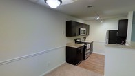 This single-floor space offers a practical and modern layout designed for efficient living. The kitchen serves as a functional centerpiece, featuring sleek dark espresso cabinetry that provides a striking contrast against the light-toned walls. Stainless steel appliances, including a gas range and an over-the-range microwave, lend a contemporary feel to the cooking area. The countertops offer adequate prep space, and the cabinetry provides ample upper storage. A decorative chair rail runs along the perimeter of the walls, adding a subtle architectural detail that elevates the overall aesthetic. The flooring transitions from light hardwood-style planks in the kitchen area to plush carpet in the adjoining living space, creating a clear distinction between zones while maintaining a cohesive design palette. Ceiling-mounted lighting fixtures, including a flush-mount light and a track light, ensure the space is well-illuminated throughout. The neutral wall tones contribute to an open, airy ambiance, making the space feel welcoming and versatile. Overall, the layout balances practicality with clean, modern design elements.