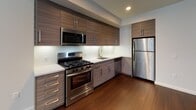 This contemporary single-floor unit offers a thoughtfully designed open-concept layout with clean lines and modern finishes throughout. The kitchen is a standout feature, outfitted with wood-grain cabinetry spanning floor to ceiling, white quartz countertops, a subway tile backsplash, and a full suite of stainless steel appliances including a gas range, built-in microwave, dishwasher, and refrigerator. Rich, dark hardwood flooring runs throughout the main living areas, adding warmth and depth to the space.

The living area is spacious and open, flowing naturally from the kitchen and filled with natural light through large floor-to-ceiling windows that offer urban views of the surrounding cityscape. The bedroom continues the hardwood flooring and benefits from the same expansive windows, creating a bright and airy feel.

The bathroom is well-appointed with light stone-look tile flooring, a soaker tub and shower combination with white subway tile surround, brushed nickel fixtures and grab bars, a floating wood-tone vanity with a white countertop, and built-in open shelving for additional storage. A large mirror completes the space.

The unit's entry features a solid dark wood door with brushed nickel hardware, setting a polished tone from the moment of arrival. Overall, the space presents a cohesive modern aesthetic with functional, high-quality finishes.