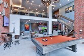 This single-floor community amenity space in Marietta, Georgia, is designed for social gathering and entertainment, blending industrial and contemporary design elements throughout. The centerpiece of the space is a dramatic double-height lounge area anchored by exposed brick walls, floor-to-ceiling windows that flood the interior with natural light, and bold pendant light fixtures that add a warm, artistic ambiance. Large-format tile flooring runs consistently throughout, unifying the various functional zones.

The lounge area is furnished with plush sofas, accent chairs, and coffee tables arranged for comfortable conversation, alongside a billiards table for recreation. A dedicated game room features a full shuffleboard table, adjustable bar-height seating, and vibrant wall art, creating a lively entertainment atmosphere. Adjacent to this is a media lounge with a multi-screen TV wall, counter-height seating, and brown leather bar stools, ideal for watching sports or other programming.

The fully equipped kitchen and bar area showcases dark cabinetry, a stainless steel refrigerator, a wet bar with sink, white subway tile backsplash, and a generous island with bar seating. Gold pendant lighting and wall-mounted televisions add a refined touch. A glass-panel door leads to an outdoor area, enhancing the sense of openness and connectivity throughout this well-appointed communal space.