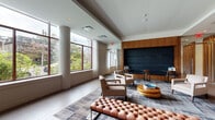 Located at 695 Atlantic St in Stamford, Connecticut, this single-floor lobby lounge is a well-appointed communal space that blends modern design with warm, inviting materials. The room features light hardwood-style flooring that transitions to large-format tile in the adjoining areas, complemented by a tray ceiling detail that adds architectural depth and visual interest overhead.

Natural light is a defining characteristic of this space, with oversized windows framed in dark wood trim lining the exterior walls, offering views of the surrounding greenery and streetscape. Sheer white curtains provide an elegant softening effect while maintaining brightness throughout the day.

The lounge area is anchored by a patterned area rug in muted blue and grey tones, upon which a curated seating arrangement is centered — including upholstered armchairs in neutral linen tones, a tufted leather bench, and a tiered circular coffee table in a warm gold finish. A wall-mounted flat-screen TV sits above a low walnut credenza flanked by tall wood cabinetry, creating a cohesive media and storage feature wall. A second accent wall showcases deep navy padded paneling set within a warm wood surround, adding a bold, sophisticated focal point. Vertical wood slat partitioning near the entry area adds additional warmth and texture to the design.