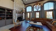 This impressive single-story communal space exudes refined elegance through its thoughtful architectural design and upscale finishes. The main living area commands attention with its soaring double-height ceilings, anchored by a dramatic floor-to-ceiling stone fireplace with a carved mantel that serves as the room's focal point. Rich hardwood flooring flows throughout, complemented by a large patterned area rug in earthy tones of teal, brown, and gold. A striking wrought-iron orb chandelier hangs overhead, casting warm light across a generously sized sectional sofa and eclectic seating arrangement that includes ornate accent chairs. Expansive arched windows line the walls, flooding the interior with abundant natural light while offering views to the outdoors. Gray-blue wainscoting and crown molding add a layer of architectural sophistication, while amber drapes provide warm contrast. A decorative console table adorned with mirrors and candelabras adds further elegance along one wall. The entryway features slate tile flooring, French doors with transom windows above, and a welcoming foyer atmosphere. The overall ambiance blends traditional and transitional design elements, creating a warm, inviting, and distinguished gathering space.