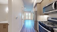 Located at 1340 S Hill St in the heart of Los Angeles' South Park district, this efficiently designed single-floor unit offers a well-planned layout comprising a living room, kitchen, bathroom, and closet. The space features light gray wood-tone flooring throughout the main areas, recessed lighting, and clean white walls that create a bright, contemporary atmosphere.

The living area is open and airy, highlighted by a set of white French doors that invite natural light and provide access to an outdoor space with greenery views — a welcome touch in an urban setting.

The kitchen is a standout feature, outfitted with light wood cabinetry accented by gold hardware, a subway tile backsplash, gray quartz countertops, and a full suite of stainless steel appliances including a gas range, over-the-range microwave, refrigerator, and dishwasher. Upper open shelving adds both function and visual appeal.

The bathroom is clean and modern, finished with large-format light tile flooring, a wood-toned vanity with gold hardware, and crisp white walls. A dedicated closet provides practical storage. A louvered closet door near the entry adds a subtle architectural detail. Smart home controls are visible near the entry, reflecting the unit's modern amenities. Overall, the space presents a polished, move-in-ready environment ideal for urban living.