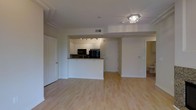 This single-floor space features an open-concept layout that seamlessly connects the living area and kitchen, creating a bright and functional environment. Light hardwood flooring extends throughout, contributing to a warm and cohesive aesthetic.

The kitchen is positioned centrally within the open layout, anchored by a peninsula with a dark granite-style countertop that provides both workspace and a natural divider between the cooking and living areas. White cabinetry lines the kitchen walls, offering ample storage, while black appliances — including a microwave and refrigerator — provide a contrasting accent. A track lighting fixture illuminates the kitchen area effectively.

The living area is generously proportioned, benefiting from a flush ceiling light fixture and a clean, unobstructed layout that offers flexible furniture arrangement. Crown molding details the ceiling perimeter, adding a subtle architectural refinement to the space.

A doorway to the right leads to an additional room, likely a bathroom, while multiple interior doors provide access to further spaces within the unit. The overall ambiance is clean, neutral, and move-in ready, with a simple, modern character throughout.