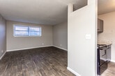 This single-floor unit offers a practical and cohesive layout with consistent wood-style flooring running throughout the living areas and bedrooms. The living room is an open, well-proportioned space featuring a ceiling fan with light fixture and a window fitted with blinds that allows natural light to fill the room. A partial wall divides the living area from the kitchen, creating a semi-open concept feel while maintaining distinct zones.

The kitchen is equipped with dark cabinetry paired with light granite-look countertops, a black electric range with coil burners, an overhead range hood, and upper cabinets providing ample storage. The flooring transitions seamlessly from the living space into the kitchen area.

The bedroom is a comfortable, naturally lit room with a window, textured ceiling, and dark hardwood-style flooring. It includes access to a walk-in closet with shelving, offering generous storage. The bathroom features a full-size white soaking tub with a tiled shower surround and chrome fixtures. Neutral gray walls are consistent throughout the unit, contributing to a clean, understated aesthetic overall.