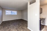 This single-floor unit offers a practical and cohesive layout with consistent wood-style flooring running throughout the living areas and bedrooms. The living room is an open, well-proportioned space featuring a ceiling fan with light fixture and a window fitted with blinds that allows natural light to fill the room. A partial wall divides the living area from the kitchen, creating a semi-open concept feel while maintaining distinct zones.

The kitchen is equipped with dark cabinetry paired with light granite-look countertops, a black electric range with coil burners, an overhead range hood, and upper cabinets providing ample storage. The flooring transitions seamlessly from the living space into the kitchen area.

The bedroom is a comfortable, naturally lit room with a window, textured ceiling, and dark hardwood-style flooring. It includes access to a walk-in closet with shelving, offering generous storage. The bathroom features a full-size white soaking tub with a tiled shower surround and chrome fixtures. Neutral gray walls are consistent throughout the unit, contributing to a clean, understated aesthetic overall.
